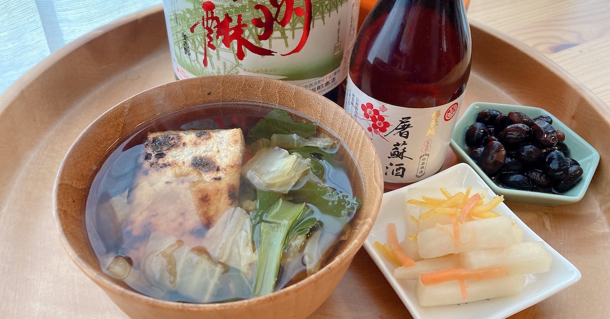 A bowl of zouni, namasu on a white plate, kuromame on a blue plate, a bottle of mirin and a bottle of otoso