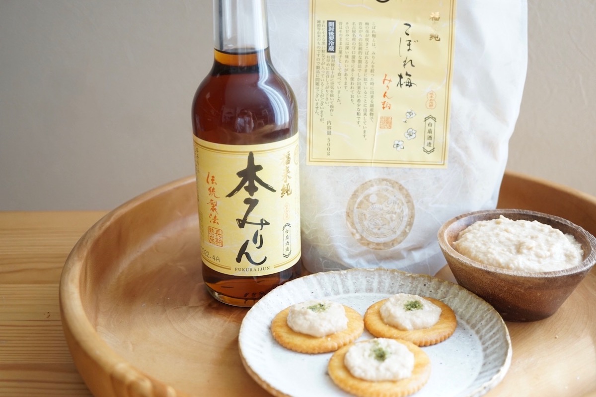 A bottle of hon mirin, a bag of kobore-ume, a bowl of dipping sauce made with kobore-ume and three crackers with dipping sauce on them placed on a wooden tray